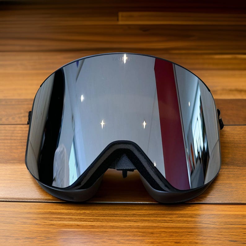 May include: Black ski goggles with a reflective, mirrored lens. The goggles have a black frame and a padded nose bridge. The lens reflects the surrounding environment, including a red and white stripe. The goggles are designed for winter sports.