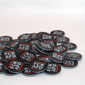 Könnte beinhalten: Ein Haufen schwarzer und weißer runder Knöpfe mit dem Text "FCK AFD" in rot und weiß gedruckt.