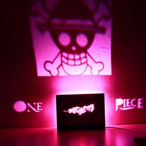 Könnte beinhalten: Eine pinkfarbene Holzbox mit den Worten "ONE PIECE" an die Wand projiziert. Das Bild projiziert auch ein Totenkopf- und gekreuzte Knochen-Logo. Die Box strahlt ein hellrosa Licht aus.