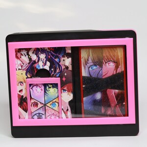 Può includere: Un espositore nero e rosa con opere d'arte in stile anime. La custodia ha una cornice rosa e contiene più pannelli con illustrazioni colorate di personaggi con grandi occhi e capelli vibranti. L'opera d'arte è dettagliata e presenta una varietà di pose ed espressioni.
