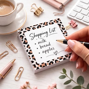 Op de afbeelding: Een notitieblok met een luipaardprintrand en de woorden "Shopping List" erop. De lijst bevat melk, brood en appels. Een hand houdt een pen vast en schrijft op het notitieblok. Andere kantoorbenodigdheden, waaronder een kopje koffie, zijn ook zichtbaar.