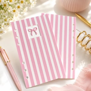 May include: Two rectangular planner inserts featuring a vertical pink and white striped pattern. The top left insert includes a small square cutout containing a pink bow icon. Each page has a series of circular punch holes along the left edge.