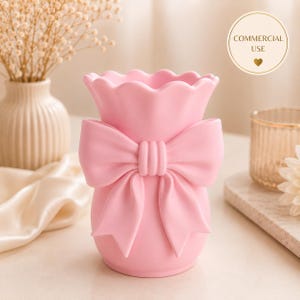 May include: A light pink ceramic vase featuring a scalloped rim and a prominent three-dimensional bow detail on the front. The vase has a smooth, matte finish and a rounded body shape, designed to hold floral arrangements or decorative items.