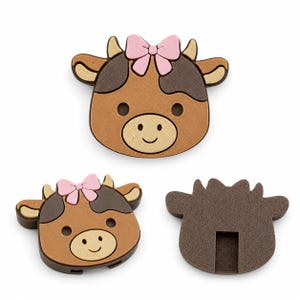 May include: Three wooden cow-shaped embellishments. Each cow has a brown and tan face with a pink bow, black eyes, and a smiling mouth. One cow is shown from the back, revealing a slot. These are likely decorative craft supplies.
