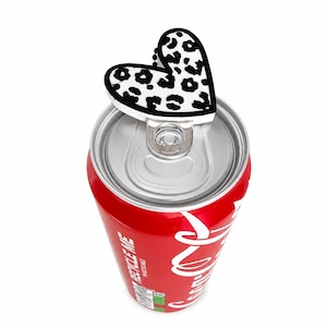 May include: A red soda can with a white logo and a black and white heart-shaped can topper. The can topper has a leopard print design. The can is open and ready to drink.