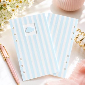 Puede incluir: Dos páginas de agenda rectangulares con un patrón de rayas verticales en azul claro y blanco. La página superior incluye un pequeño recorte cuadrado que contiene un icono de nube azul claro. Cada página tiene perforaciones circulares a la izquierda.