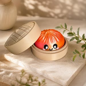 May include: A round, orange-colored dumpling with a hand-painted smiling face featuring black eyes and pink cheeks, resting inside a light beige, circular bamboo steamer basket with a matching lid tilted to the side, showcasing a grid pattern.