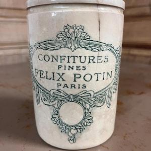 Peut inclure: Pot en céramique ancien avec couvercle. Le pot est blanc cassé avec une étiquette décorative verte qui indique "CONFITURES FINES FELIX POTIN PARIS". Le pot a une forme cylindrique et une fissure sur le côté.