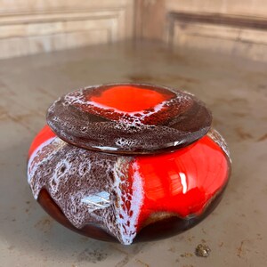 Peut inclure: Un pot en céramique avec un couvercle. Le pot est rond avec un glaçage rouge et marron brillant. Le couvercle est marron avec un centre rouge et des accents blancs. Le pot est probablement utilisé pour le stockage ou la décoration.