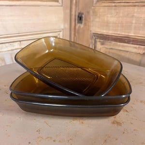 Peut inclure: Trois plats à four en verre ambré empilés. Chaque plat a une forme rectangulaire avec des coins arrondis et un bord foncé. Le plat supérieur a un motif nervuré sur le fond. Les plats sont en verre.