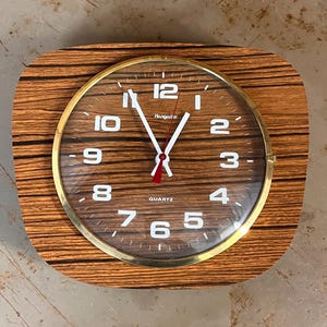 Può includere: Un orologio al quarzo vintage con cornice in legno e lunetta color oro. Il quadrante dell'orologio ha numeri e lancette bianche, con una lancetta dei secondi rossa. L'orologio indica "Quartz" in basso. Il marchio "Hongate" è stampato sul quadrante.