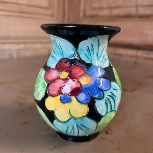 Könnte beinhalten: Kleine, handbemalte Keramikvase mit schwarzem Boden und Rand. Die Vase zeigt ein Blumenmuster mit roten, blauen und gelben Blüten, umgeben von grünen Blättern, vor schwarzem Hintergrund. Die Vase ist etwa 13 cm hoch.