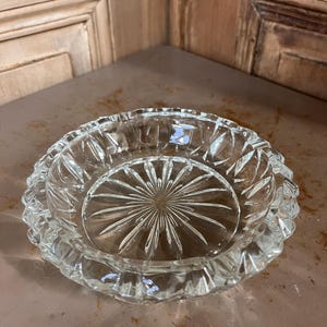 Peut inclure: Un cendrier rond en verre transparent avec un motif en étoile au centre et un bord festonné. Le cendrier est en verre épais et repose sur une surface brun clair. Le design est vintage.