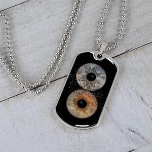 Könnte beinhalten: Halskette mit silberfarbenem Anhänger in Form einer Erkennungsmarke mit zwei detaillierten Augeniris-Designs. Die Iris ist in Blau-, Braun- und Schwarztönen gehalten und vor einem schwarzen Hintergrund mit weißen Sprenkeln platziert. Die Halskette hat eine Kugelkette.
