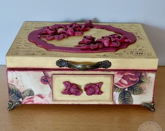Può includere: Una scatola decorativa in legno con base color crema e cornice bordeaux. La scatola presenta un motivo floreale con rose bordeaux in rilievo e una maniglia decorativa. I lati sono adornati da illustrazioni di rose e dettagli angolari ornati.