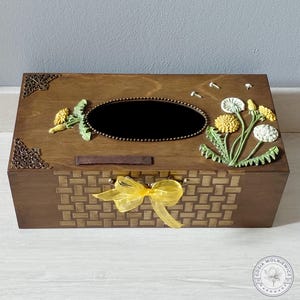Pode incluir: Uma caixa de madeira com um padrão tecido na frente e uma fita amarela. A caixa apresenta detalhes decorativos nos cantos e uma abertura oval central. Flores de dente-de-leão esculpidas no topo. A caixa tem 25 cm de comprimento, 15 cm de largura e 10 cm de altura.