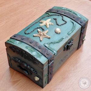 May include: A rectangular wooden chest with a teal finish, featuring dark rusted metal bands, a metal latch, and a handle. The lid is decorated with two beige starfish, a small seashell, and a thin metal chain. The text PARIS 1923 is visible.