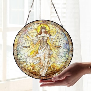 May include: Round stained glass panel featuring the Libra zodiac sign. The artwork depicts a robed figure holding scales, set against a backdrop of warm yellow and gold tones. The panel is framed in black and hangs from a chain.