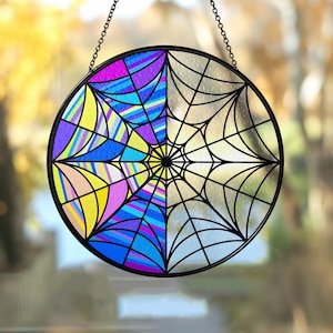 May include: A round stained glass window hanging with a black metal frame and chain. The left side features a colorful geometric design in shades of yellow, pink, blue, and purple. The right side has a black spiderweb design.