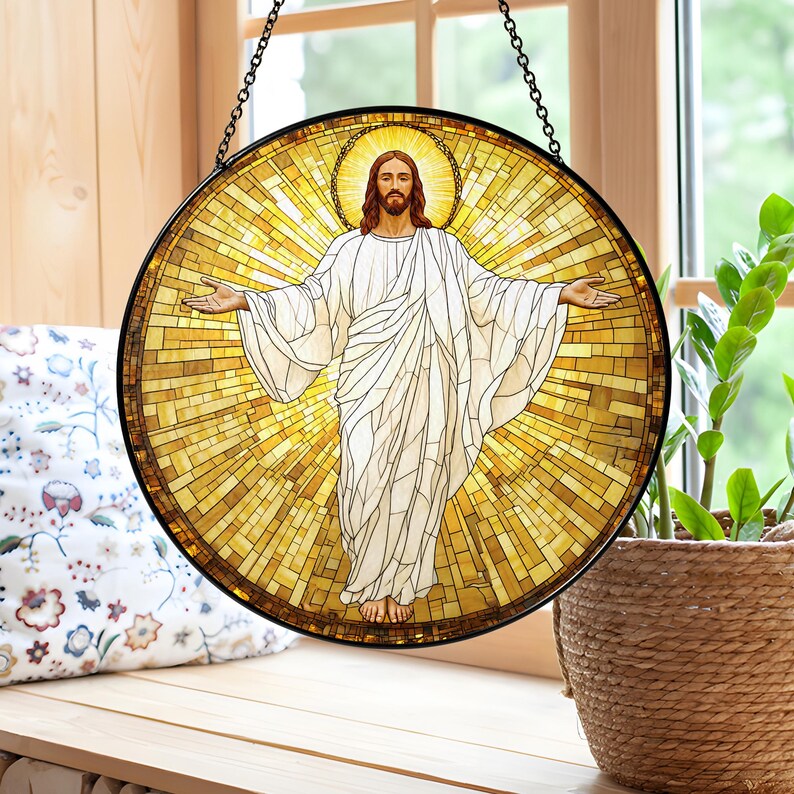 May include: Round stained glass window art depicting a figure with outstretched arms, surrounded by a golden halo and background. The figure is wearing a white robe. The window is framed in black and hangs from a chain.