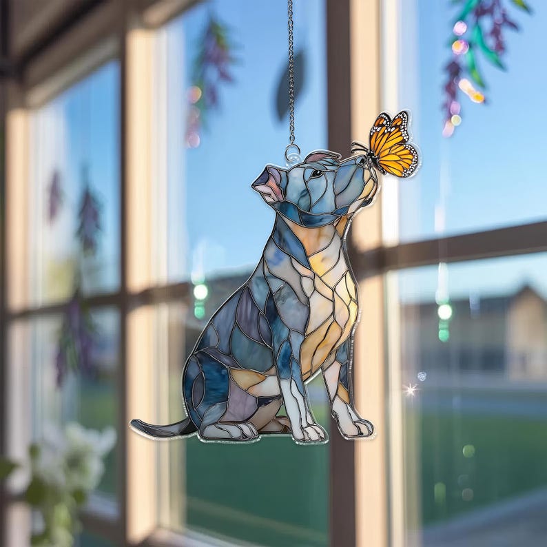 May include: Stained glass dog suncatcher in shades of blue, yellow, and beige. The dog is sitting and looking up at a butterfly with orange and black wings. The suncatcher hangs from a silver chain, perfect for window decor.