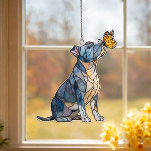 May include: A stained glass dog figurine in shades of blue, purple, and yellow, with a butterfly perched on its nose. The dog is sitting, and the figurine hangs from a silver chain. The background shows a window with an autumn scene.