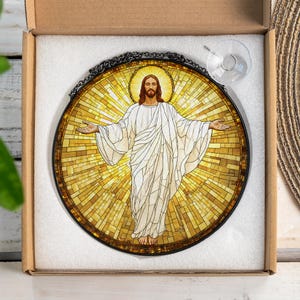 May include: Round stained glass window art featuring a depiction of Jesus with outstretched arms. The artwork has a mosaic-style background in shades of yellow and gold, with a black metal frame and chain for hanging. A suction cup is attached.