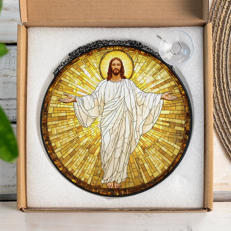 May include: Round stained glass window art featuring a depiction of Jesus with outstretched arms. The artwork has a mosaic-style background in shades of yellow and gold, with a black metal frame and chain for hanging. A suction cup is attached.