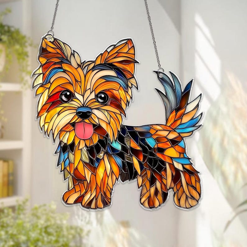 Op de afbeelding: Gebrandschilderd glas van een Yorkshire Terrier met een vrolijke uitdrukking, roze tong en blauwe neus. De vacht van de hond is een moza&iuml;ek van oranje, gele, bruine en blauwe glasstukken. Het hangt aan een zilveren ketting.