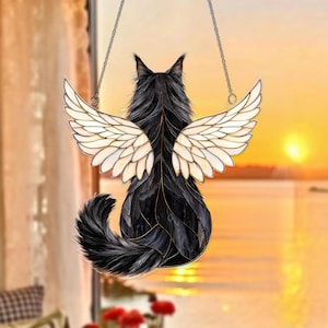 May include: A stained glass suncatcher depicting a black cat viewed from behind with large, cream-colored feathered wings. The cat has a long, fluffy tail and pointed ears. The piece is suspended by two thin metal chains against a sunset background.