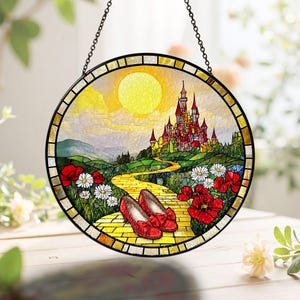 May include: Round stained glass panel featuring a yellow brick road leading to a castle under a bright sun. Red shoes with bows sit on the road, surrounded by red poppies and white daisies. The panel is framed in black and hangs from a chain.