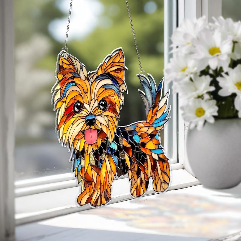 Op de afbeelding: Gebrandschilderde Yorkshire Terrier hond zonnevanger met een moza&iuml;ek ontwerp. De hond heeft een kleurrijk patroon van oranje, geel, blauw en zwart. Hij heeft een roze tong en hangt aan een ketting. De zonnevanger wordt getoond bij een raam.