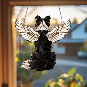 May include: A decorative window hanging featuring a black and white dog with angel wings. The dog is depicted from the back, with its wings spread. The wings are white with detailed feathering, and the dog's fur is black and white.