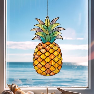 May include: A stained glass pineapple suncatcher with a chain for hanging. The pineapple features a yellow and orange body with green and yellow leaves. The background shows a blue sky and ocean.