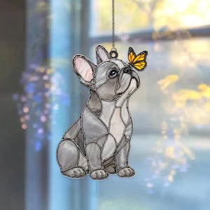 May include: A stained glass suncatcher featuring a grey French Bulldog sitting and looking up at a small orange and black monarch butterfly perched on its nose. The piece is constructed with dark metal lead lines and hangs from a silver chain.