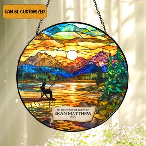 May include: A circular stained glass suncatcher featuring a landscape of mountains, a lake, and a person fishing from a dock at sunset. The text reads CAN BE CUSTOMIZED and IN LOVING MEMORY OF ERAN MATTHEW 2025. The piece has a black metal frame.