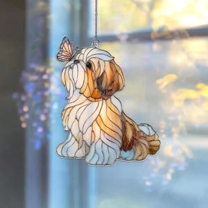 May include: A decorative stained glass Shih Tzu dog with a butterfly perched on its head. The dog is primarily white, brown, and tan, with a silver chain for hanging. The butterfly has orange, yellow, and purple accents. A charming window decoration.