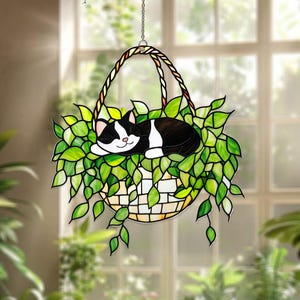 May include: A stained glass hanging decoration featuring a black and white cat sleeping in a basket of green leaves. The basket has a woven handle and is suspended by a chain. The background is a blurred view of a window and plants.