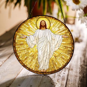 May include: Round stained glass window depicting a figure with outstretched arms, wearing a white robe, and a golden halo. The background is a mosaic of yellow and gold tiles. A black chain is attached to the top for hanging.