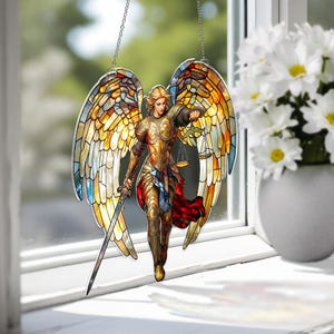 May include: Stained glass window hanging featuring an angel with outstretched wings, a sword, and scales. The angel is depicted in gold and bronze armor, with colorful wings in shades of yellow, orange, and blue. The window hanging is suspended by a silver chain.