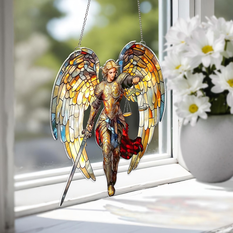May include: Stained glass window hanging featuring an angel with outstretched wings, a sword, and scales. The angel is depicted in gold and bronze armor, with colorful wings in shades of yellow, orange, and blue. The window hanging is suspended by a silver chain.