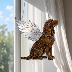 May include: A stained glass suncatcher depicting a brown spaniel dog in a sitting position with large, translucent white wings. The dog features detailed dark brown fur textures and is suspended by two thin metal chains for hanging display.