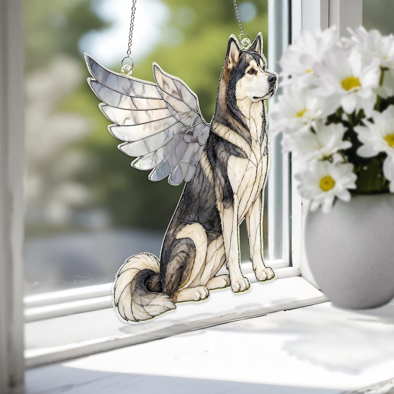 May include: A stained glass suncatcher depicting a sitting husky dog with large, translucent wings. The dog features black, grey, and white segments with dark outlines. The wings are composed of clear, textured glass panels. It hangs by a metal chain.