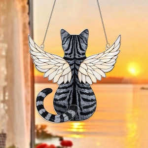 May include: A decorative cat-shaped window hanging. The cat is depicted from the back with gray and black stripes, and white angel wings. The cat is suspended by a silver chain. The background shows a sunset over the water.