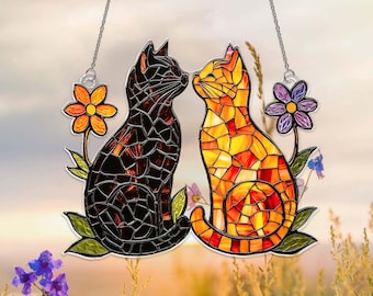 Puede incluir: Atrapasoles de gato de vidriera con dos gatos en un abrazo amoroso. Un gato es negro, el otro es naranja y amarillo. Cada gato está rodeado de flores y hojas, con una cadena para colgar.