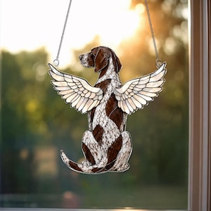 May include: A stained glass dog with angel wings, featuring a brown and white spotted pattern. The dog is sitting, with its wings spread. A silver chain is attached for hanging. The background is a blurred view of a window.