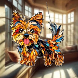 Op de afbeelding: Een gebrandschilderd glazen Yorkshire Terrier hond zonnevanger. De hond is afgebeeld in een moza&iuml;ekstijl met levendige kleuren, waaronder oranje, geel, blauw en rood. Het heeft een roze tong en wordt opgehangen door een zilveren ketting. De achtergrond is wazig.
