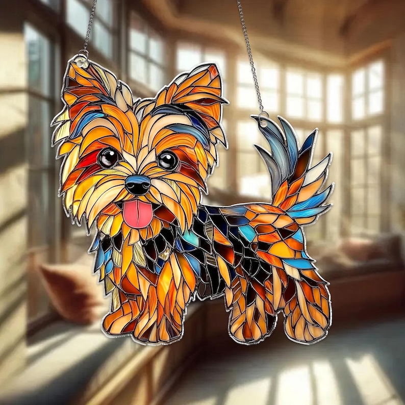 Op de afbeelding: Een gebrandschilderd glazen Yorkshire Terrier hond zonnevanger. De hond is afgebeeld in een moza&iuml;ekstijl met levendige kleuren, waaronder oranje, geel, blauw en rood. Het heeft een roze tong en wordt opgehangen door een zilveren ketting. De achtergrond is wazig.