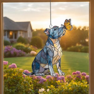 May include: Stained glass window hanging featuring a seated dog with a butterfly on its nose. The dog is primarily blue with yellow and white accents. The butterfly is orange and black. The background shows a house and garden.