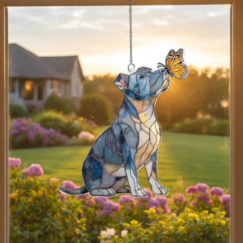 May include: Stained glass window hanging featuring a seated dog with a butterfly on its nose. The dog is primarily blue with yellow and white accents. The butterfly is orange and black. The background shows a house and garden.